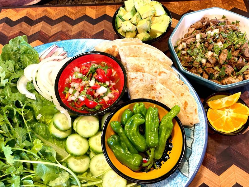 A colorful taco spread with salsa, avocado, shishito peppers, greens and warm tortillas