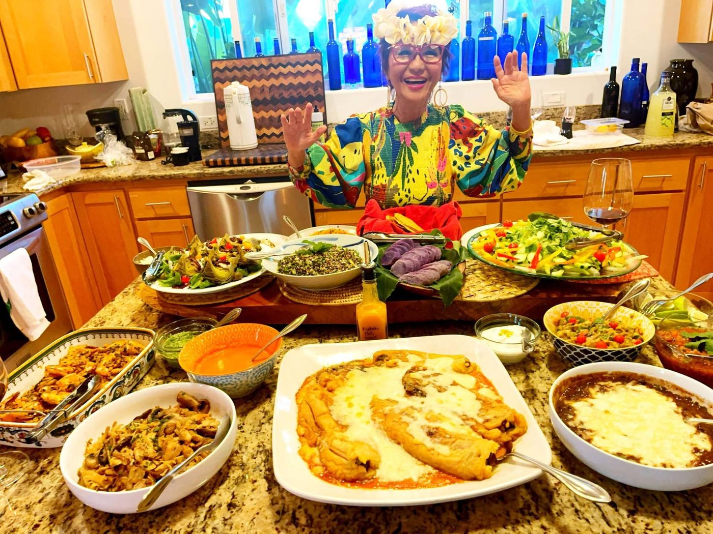Olelo paʻa in a flower crown, laughing with her hands raised behind a kitchen island full of dishes she has cooked